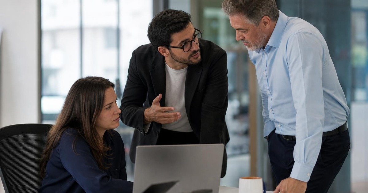 Three adult professionals in a modern office discussing a stalled project while one colleague calmly seeks a decision from a senior teammate.
