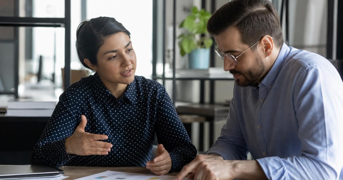 Two adult colleagues in a modern office having a brief progress check-in over a shared project plan, with calm collaborative body language.